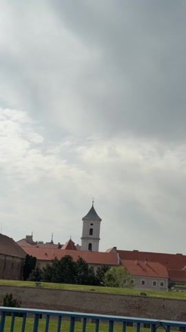 The camera records the exterior view of what appears to be an old, possibly religious, complex. In the foreground, a blue railing is visible. Behind it is a short stone wall topped with grass.  The complex consists of various buildings with red-tiled roofs. The tallest structure is a white tower with a pointed spire topped by a cross. The sky is overcast with a mix of white and grey clouds.  No people are visible. The camera is mostly static, with a slight horizontal movement.