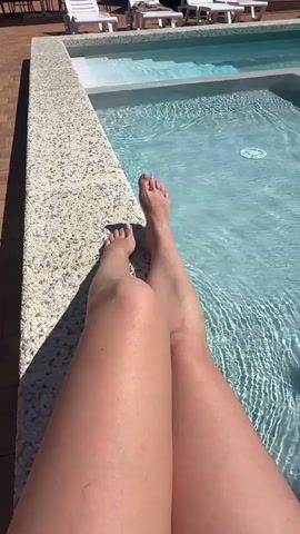 A first-person perspective video shows a person's legs and feet extended over the edge of a swimming pool. The person's legs are tan and bare, with the toes of their left foot visible near the water's surface. The pool is filled with clear blue water, with sunlight creating rippling patterns on the surface. In the background, lounge chairs and wooden decking are visible. The video is shot during the day, with bright sunlight casting shadows.