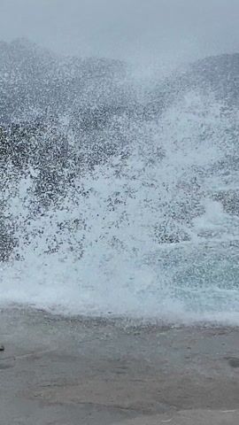 The video depicts a turbulent sea scene. Waves crash against rocky cliffs and a concrete platform, creating white foam. The water color is a mix of dark blue and turquoise. In the background, there are hazy mountains and a muted sky suggesting overcast conditions. The camera perspective is from a relatively low vantage point on the concrete surface, capturing the dynamic interaction of the waves with the shore.
