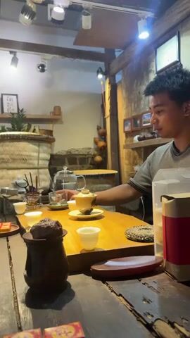A young man with short, dark hair and a grey t-shirt is sitting at a wooden table, participating in a tea ceremony. He is holding a lid to a small teapot with a brown and gold pattern. In front of him, on the table, are several small white tea cups and a larger, ornate teapot. There is also a dark brown, textured lump of what appears to be tea, placed on a small wooden stand. The table is set up with various tea-related implements. The background shows a rustic room with wooden walls and shelves adorned with decorative items, including a woven basket and some gourds. The lighting is warm and somewhat dim, with spotlights illuminating parts of the scene. The man's gaze is directed towards the teapot.