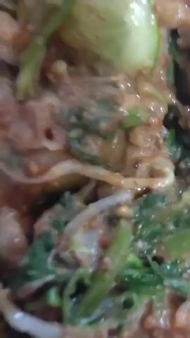 The video displays a close-up view of a dish, likely a salad or stir-fry, served on paper. The dish contains a mixture of vegetables such as leafy greens and bean sprouts, along with what appears to be pieces of protein and a brown sauce. A spoon is visible, resting on the paper beside the food. The background suggests an indoor setting, possibly a table covered with a patterned cloth. The camera angle is overhead, providing a top-down perspective of the meal.