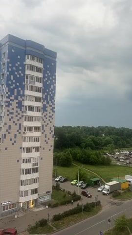 The video is a vertical, overhead shot of a modern residential building and its surroundings. The building, predominantly beige with blue accents forming a pixelated pattern on its side, has multiple floors with balconies and windows. In front of the building, a paved area serves as a walkway and a small parking lot. Several cars are parked, including a white truck. A road runs alongside the parking area. Beyond the building and parking lot, a patch of green grass leads to a dense expanse of trees that stretches to the horizon. The sky is overcast with dark, ominous clouds. The scene appears to be an urban or suburban environment, possibly on a cloudy or stormy day. There are no visible people performing any significant actions in the short clip.