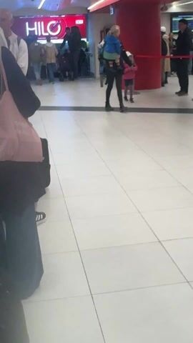 The camera is still, offering a slightly side-angled view from the ground level of what seems to be an airport or train station. The floor is white with square tiles separated by black lines. To the left, parts of a person wearing a dark dress or jacket and blue pants are visible, along with a pink bag with white lettering. 

Moving towards the center of the frame, a woman dressed in dark clothing is carrying a small child wearing a blue jacket and green pants. The child is partially facing the woman, appearing to be held close. A red column is visible in the background, along with a sign that reads "HILO" in a red and white font.

In the background, a group of people is gathered near what appears to be a ticketing area, with a man standing in front of a stroller. Another person is walking to the right of the frame. The lighting in the area is bright and artificial.