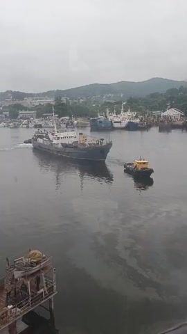 The video shows a harbor scene with several boats. The main focus is on a large ship being towed by a smaller yellow tugboat. The large ship is dark gray and appears to be moving through the water. The tugboat is positioned behind and to the side of the large ship, with a tow line connecting them. In the background, there are other ships docked, and buildings and hills are visible on the shore. The sky is overcast, suggesting a cloudy or possibly rainy day. In the foreground, there is a structure that looks like a pier or a platform with some equipment on it, and two figures can be seen on the structure.