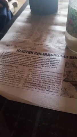 The video shows a close-up view of a newspaper article, with the camera moving slightly. The article appears to be in a foreign language, likely Kazakh, given the Cyrillic script. The headline translates to something about "Winter is coming, what is the readiness of the country." The article discusses infrastructure and mentions cities and distances, suggesting it's related to the country's preparedness for winter. The newspaper is lying on a dark surface, possibly a table, and there's a blurry object in the background, which could be a drink or a container. The lighting is dim, creating a somewhat muted atmosphere. There are no people clearly visible in the frame.