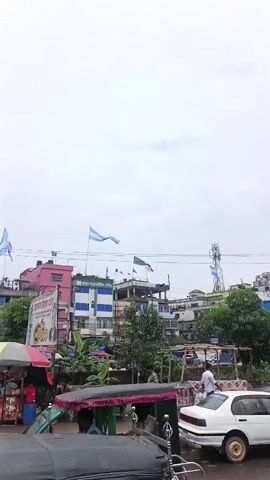 The video shows a view of a cityscape on a cloudy day. In the foreground, a portion of a vehicle is visible, suggesting the video is being filmed from inside a car or similar mode of transport. The middle ground is dominated by buildings of varying heights and colors, some adorned with flags, including what appear to be Bangladeshi flags and other national flags. There are also a few people walking on the street. Overhead, a complex network of power lines crisscrosses the sky, and a cell tower is visible. The background features more buildings and trees, further contributing to the urban landscape. The overall impression is a bustling, populated area.