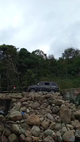 The video shows an outdoor scene with a dark SUV parked on a makeshift bridge over a small stream. The bridge is constructed over large rocks. A person standing among the rocks takes a photo of the SUV with a phone. In the background, lush green trees and an overcast sky are visible. Later, the camera pans to reveal a white van parked on a gravel road nearby. The surroundings suggest a mountainous or hilly area.
