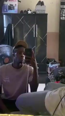 A young man stands in front of a mirror, holding a phone up to take a selfie or video. He is wearing a light gray t-shirt and a tan baseball cap. Behind him is a black wardrobe with a geometric design in white or silver, a fan, and other items scattered around the room. The lighting is soft, and the focus is on the man in the mirror.