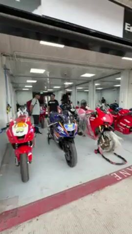 The video shows a pit lane at a race track. Several motorcycles, mostly red, are parked inside a pit garage. People are seen standing around.  The garage has a sign indicating a partnership between Pertamina Enduro and Ducati Indonesia. The camera pans to show the pit lane, which is a gray concrete surface with a red and yellow line. The pit garages are closed with roller shutters.  In the distance, a green landscape can be seen beyond the track. The sky is overcast.