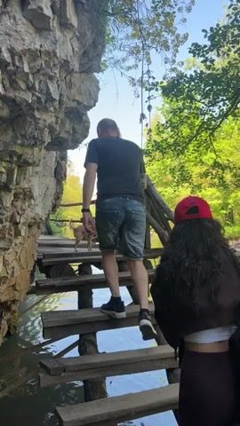 The video shows a group of people walking on a wooden staircase bridge. The bridge is built next to a rock cliff and passes over water. The first person in the group is a man wearing a black t-shirt and denim shorts. He is followed by a woman with curly hair wearing a red baseball cap, brown pants, and a black backpack. A small dog is also seen following the man. The environment includes trees and greenery, and the sky is visible in the background.