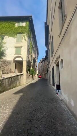 The video shows a narrow cobblestone street in a town. The buildings on either side are old and made of stone or stucco. There are green shutters on some of the windows, and ivy grows on one building. In the distance, a red car is visible. The street is empty except for a woman in the foreground who is filming herself. She is wearing a white shirt and sunglasses. There are also some trash bins and a bicycle visible. The sky is blue with some clouds.