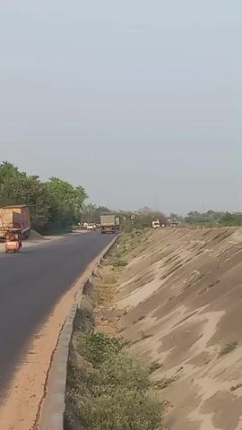 A quick shot from a moving vehicle captures a scene on a two-lane highway. The road is black asphalt with a dirt shoulder. On the right is a dirt embankment covered with sparse vegetation. A few trucks and other vehicles travel along the road in the distance.  Later, the camera focuses briefly on a small roadside stand and a vacant dirt lot.  The scene ends with a view of a fence and tree through the lens of the sun.  The sky is overcast.