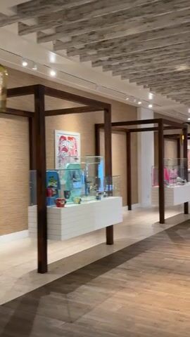 The video shows a wide shot of an indoor walkway, possibly a mall or a resort. The walkway has a wooden floor and a textured ceiling with exposed beams. On the left side of the walkway, there are display cases with items inside, each housed in a wooden frame structure. The display cases are white and topped with glass enclosures. Beyond the display cases, the wall is textured like cork or grasscloth. Further down the walkway, there are tables and chairs set up, suggesting a dining area or lounge. On the right side of the walkway, people are walking towards the camera, some of whom are looking at the display cases. The lighting is warm and ambient, with pendant lights hanging from the ceiling. The overall atmosphere appears to be calm and inviting.