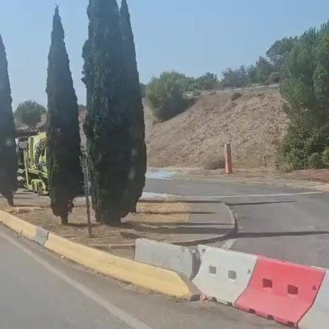 The video pans across a scene of vehicle wreckage. A flatbed truck, also yellow, appears to have been carrying other vehicles. One of these vehicles, a yellow emergency vehicle, is severely damaged and appears to be on fire or has recently been on fire. Its front end is crushed, and debris is scattered around it. The flatbed truck itself seems intact but is also positioned in a way that suggests it may have been involved in the incident. The scene is outdoors, with a paved area, dry grass, and tall, slender trees. A hilly, vegetated area is visible in the background, along with a lamppost. There are no people clearly visible in the video.