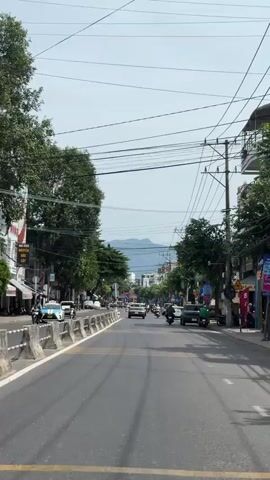 A vertical, eye-level shot shows a street scene in what appears to be a city, likely in Southeast Asia, on an overcast day. The road is paved and marked with yellow and white lines. A median barrier with concrete sections and some foliage runs along the left side of the road. Buildings with signs and windows line the street on both sides, interspersed with trees and utility poles. Numerous overhead electrical wires crisscross the sky. Traffic includes cars, motorcycles, and a delivery vehicle. On the left side of the road, several motorcycles are parked along the sidewalk. A few people are visible, mostly on motorcycles. The background reveals more buildings and distant hills or mountains under a pale sky. The camera appears to be moving forward, suggesting a ride-along perspective.