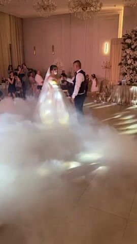 A wedding reception is captured, focusing on the bride and groom dancing in the center of the room amidst a cloud of fog. The bride wears a white off-the-shoulder wedding dress with a veil. The groom is in a white shirt, black vest, and dark pants. They are holding hands and appear to be in the middle of their first dance. Surrounding them, guests are seated at tables, some standing and observing the couple. The room is decorated with chandeliers, floral arrangements, and ambient lighting, including a projector casting circular patterns of light on the ceiling. A photographer is visible in the foreground on the left, holding a camera, and a professional lighting setup is positioned on the right. The overall atmosphere suggests a celebratory and romantic occasion.