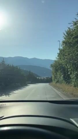 The video shows a view from inside a vehicle, looking out through the windshield. The sun is shining brightly in the upper left corner of the frame, creating lens flare. A two-lane paved road curves to the right, leading into a mountainous landscape with layers of blue hills in the distance. On the right side of the road, there are tall green trees and a lamppost. On the left side of the road, there are more trees and a few scattered buildings or structures. As the video progresses, a motorcyclist on a dark-colored motorcycle comes into view, riding away from the camera down the road. The motorcyclist is wearing a helmet. The steering wheel of the vehicle is visible in the foreground. The overall scene depicts a scenic drive on a sunny day.