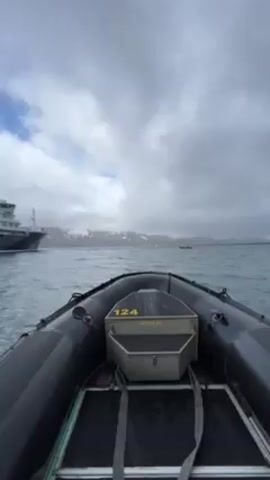 The video shows a view from a small, black inflatable boat, numbered "124", moving through choppy water. The sky is overcast with gray clouds. In the distance, there are snow-capped mountains and land. A larger vessel is visible to the left side of the frame in the beginning. The boat is pointed towards the distant land. There are a few other small boats visible in the water. The boat seems to be moving at a moderate pace.