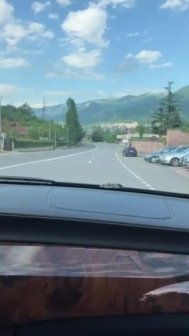 The video captures a scenic drive through Ijevan, Armenia. The perspective is from inside a car, looking out through the windshield. The road ahead curves gently, bordered by lush greenery on the left and a stone wall and buildings on the right. In the distance, majestic mountains rise, their slopes covered in trees, under a bright blue sky dotted with fluffy white clouds.

The time of day appears to be late morning or early afternoon, judging by the bright sunlight and clear skies, suggesting pleasant weather. The activity is a car journey, with the vehicle moving forward along the paved road.

Notable details include the dashboard of the car in the foreground, which features a polished wood-grain trim and black leather accents. Several cars are visible on the road and parked along the side. One car, a dark sedan, is parked on the right side of the road. Further down, another car, possibly a silver sedan, is partially visible.

There are some visible signs and structures along the road. A traffic light with red and white markings is positioned above a red directional sign with white arrows. There is also a building with a flat roof and large windows, appearing to be a commercial establishment. A flagpole with a flag is visible in front of this building. Further along the road, there are other buildings and what appears to be a small market stall. The overall impression is one of a peaceful, scenic drive through a town nestled amongst mountains.