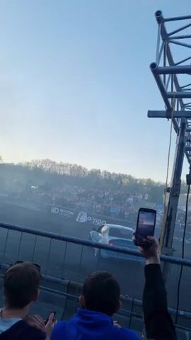 The video shows a light blue car drifting at a race track. Spectators watch and take pictures with their phones from behind a fence. The scene is filled with smoke, likely from the car’s tires. The background features trees and a clear sky. There is a metal structure to the right of the frame. Some spectators are visible in the foreground.