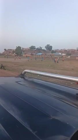 The video shows a soccer game in progress on a dirt field. The camera is positioned on the roof of a black vehicle, partially visible in the foreground. People in different colored shirts, presumably the soccer players, are spread across the field actively playing. Buildings and trees form the backdrop, suggesting a rural or suburban setting. Other people are observing the game from the side of the field. A person's hand can be seen intermittently holding onto the roof rack of the vehicle, possibly to maintain stability. The sky is overcast, indicating either early morning or late afternoon.