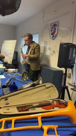 A man wearing glasses, a brown tweed jacket, a white shirt, and green pants is playing a saxophone. He is standing in a room with a blue table in the foreground covered with various musical instruments, including a violin in an open case, a trombone, a clarinet, and a trumpet. A whiteboard with musical notes is visible on the left. A speaker is on a stand to the right. The wall behind the man has a crest and musical notes decoration. There are two children sitting at the table on the left. The video pans slightly from right to left.