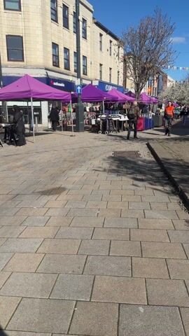 The video captures a bright, sunny day at an outdoor market situated on a paved street. Numerous market stalls are set up on the left side of the street, each covered by a distinct purple gazebo or tent. These stalls display various goods, including what appear to be handbags and other accessories, arranged on tables and racks. Buildings with multiple stories line the street behind the stalls; one building clearly shows a "Nationwide" sign, and another has "CORA" visible on its facade.

The sky is a clear, vibrant blue, and the sun casts strong, defined shadows on the light grey paved ground. Colorful bunting, consisting of small flags, is strung across the street in the distance, adding a festive touch to the scene. The trees lining the street are bare, suggesting the season might be late winter or early spring.

Several people can be seen moving through the market. Early in the video, a man wearing an orange and black high-visibility jacket walks from the right side of the frame towards the left, carrying a dark object. Later, a woman dressed in light-colored clothing walks from left to right, pulling a red rolling suitcase behind her. Other individuals are visible further down the street, some appearing to be browsing the items at the market stalls, contributing to a lively atmosphere.