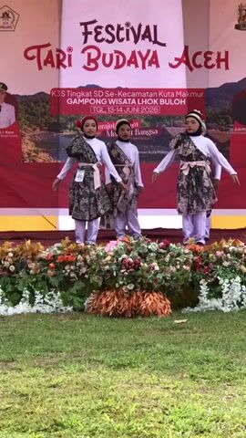 Four young girls in traditional attire are performing a dance on a stage decorated with flowers. The stage backdrop features text that appears to be in Indonesian, reading "FESTIVAL TARI BUDAYA ACEH" and other event details. The girls are wearing matching patterned dresses over light-colored pants and headscarves, with sashes tied around their waists. They are performing synchronized movements with their arms and hands. The event seems to be outdoors, with green grass visible in the foreground and trees in the background.