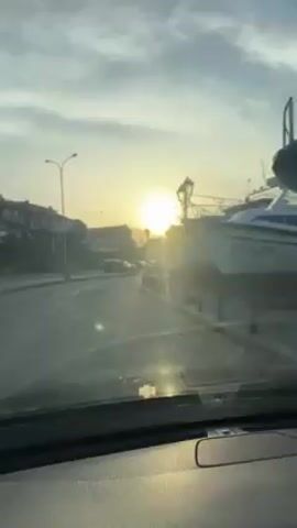 The video shows a view from inside a car driving on a road in what appears to be a coastal town or city. The sun is low in the sky, creating a bright glare. Parked along the side of the road are several boats on land, presumably for maintenance or storage. A few vintage-style streetlights line the edge of the road. In the background, a line of parked cars is visible, and some buildings are faintly outlined against the sky. The windshield of the car is a bit dirty.