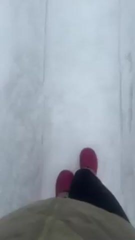 A person wearing pink boots, black leggings, and a tan jacket walks in a narrow path dug out of snow. The walls of the path are tall and white. The snow inside the path is also white and appears deep. The camera angle is from the person's perspective, looking down at their feet as they walk. The video is shaky, indicating handheld recording.