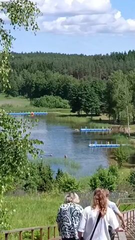 The video begins with a view of a lake surrounded by lush green trees under a partly cloudy sky. Several floating blue platforms are visible in the water, some with people swimming nearby. On the right side of the screen, a wooden walkway leads down a grassy slope. Three people, two women and one person whose gender is unclear, are walking down this wooden walkway. The woman in the foreground on the left has blonde hair and is wearing a patterned shirt. The woman in the middle has long, reddish-brown hair and is wearing a white t-shirt and a dark skirt, carrying a black bag. The third person is partially obscured. The walkway continues to curve around the slope, with a mown grassy area to its right. The background shows a dense forest and some scattered buildings.