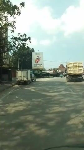 A video shows a view from inside a vehicle moving along a road. A large billboard with an image of a mango is visible on the left side of the road. Several vehicles, including trucks and a car, are also on the road. The sky is cloudy.