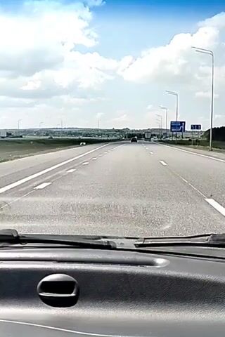 The video shows a view from inside a car as it drives on a highway. The dashboard and windshield wipers are visible in the foreground. The road ahead has multiple lanes and is lined with streetlights. There are other vehicles, including cars and trucks, on the highway. A blue sign with white text and arrows is visible on the right side of the road, indicating directions to Moscow and Kumatori. The sky is blue with white clouds. The landscape on either side of the highway appears to be rural, with fields and trees.