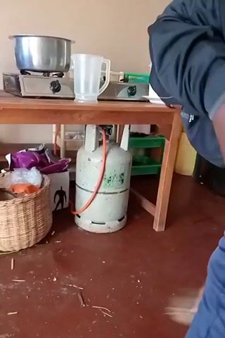 Here is a concise description of the video:

A person is sweeping a floor with a traditional broom. The floor is red, and there are scattered pieces of debris on it. The person is wearing a black jacket and blue pants, and is using red flip-flops. A wooden table, a wicker basket, a cooking pot, and a green sack are also visible in the room.
