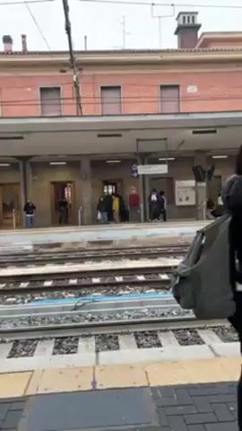 The video shows a train platform on a slightly overcast day. People are waiting with luggage, including rolling suitcases, backpacks, and shopping bags. The platform has tiled flooring and is covered by a canopy. Railway tracks run parallel to the platform. A brick building with windows stands across the tracks. There are signposts indicating track numbers. Some people are standing alone, while others are in small groups, some appear to be talking. One person rolls a suitcase, others are looking at their phones.
