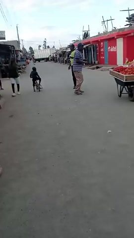 The video shows a street scene in what appears to be a village or small town. A young child is riding a small bicycle down the road. Several adults are standing or walking nearby. There are shops or stalls lining both sides of the street. The road is paved, and the sky is overcast. The camera follows the child on the bicycle and occasionally pans to show the surrounding area.