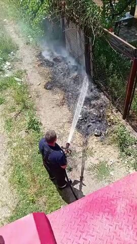 An overhead shot shows a firefighter in a blue uniform and black suspenders spraying water from a hose onto smoldering debris near a metal fence. The debris consists of burnt material, ash, and some refuse, with plumes of white smoke rising. The ground is a mixture of dirt and sparse grass. A red textured surface, likely part of a fire truck, is visible in the lower right corner of the frame. The background shows green foliage and trees.