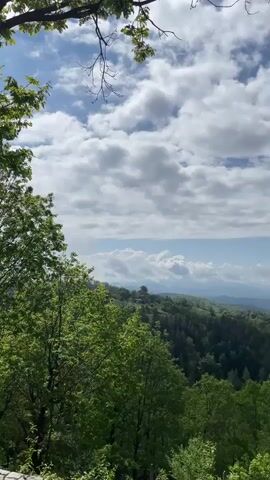 The video showcases a scenic, mountainous landscape viewed from a slightly elevated perspective. Lush green trees dominate the foreground, their leaves vibrant under the daylight. In the distance, rolling hills covered with dense forests stretch towards the horizon. The sky above is a mix of blue and white, with puffy clouds scattered across the expanse. A stone ledge or wall is visible in the frame. The camera moves from left to right slightly, panning across the view.
