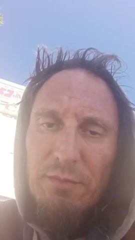 The video is a close-up shot of a middle-aged man. He has dark hair that is unkempt and a short beard. He is wearing a grey hooded sweatshirt. The man is looking directly at the camera and speaking. In the background, there is a light-colored building with faded text and a blue sky.