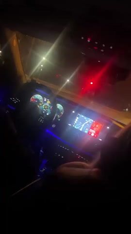 The video shows the interior of a car at night, with the driver's side visible and the dashboard illuminated. The steering wheel and digital displays are lit with blue and green lights. A person's hands are on the steering wheel. The car appears to be stopped, possibly at a traffic light, as a red light is visible in the background, reflecting off the windshield and dashboard. The passenger side of the car is mostly in shadow, but a person's head and shoulders are visible, facing forward. The overall atmosphere is dark, with the car's interior lights and the external red light providing the primary illumination.