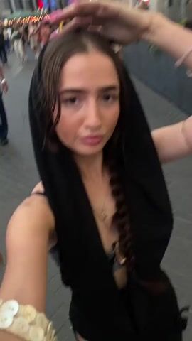 The video shows a young woman from the chest up, taking a selfie as she walks outdoors. She is wearing a black head covering that drapes over her shoulders and down her chest, a black tank top, and a gold necklace. Her hair is braided into a single plait that hangs down her back. On her left wrist, she wears a bracelet made of numerous flat, round, off-white discs. She is walking on a paved walkway, with blurred figures and lights visible in the background, suggesting a public space or event. Her right hand is raised towards her head, possibly adjusting her head covering or posing for the camera. The video is shot from a low angle, looking up at the woman.