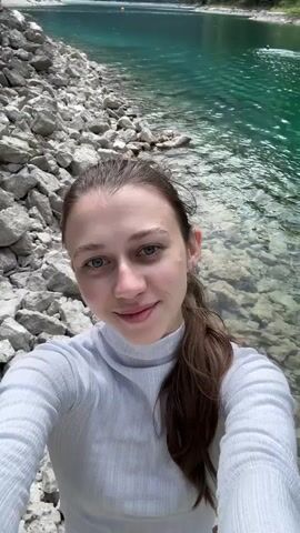 A young woman with brown hair pulled into a ponytail is taking a selfie at the edge of a clear, turquoise lake. She is wearing a white turtleneck shirt. The lake is surrounded by rocky shores and lush green forests, with a majestic mountain range in the background. The water is calm, and a few people can be seen swimming in the distance. The sky is clear and blue. The video appears to be a tourist video or a personal vlog.