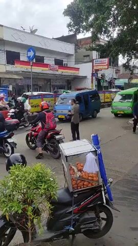 The video shows a street scene in what appears to be a Southeast Asian city. There are several motorbikes, one with a glass display case attached to the back, filled with round, orange food items. The motorbike is parked near a small green bush.  A man in a blue shirt walks past a blue van. Another man on a red motorbike with a red backpack waits at the intersection. Behind him is an "Alfamart" convenience store.  A green public transportation van is also visible. Other motorbikes pass by. Another man walks across the street wearing a long-sleeved shirt, dark pants, and a baseball cap.  The sky is overcast.  Traffic appears moderate.