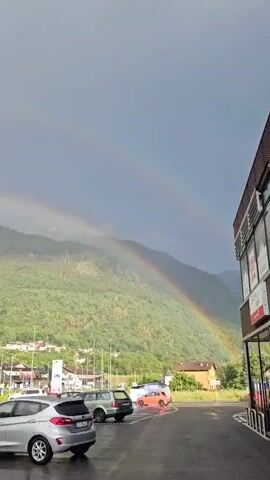 A vertical shot captures a cloudy, overcast sky with two rainbows visible arching over a green, tree-covered mountainside. Below the mountain, a small town with buildings and streets can be seen. In the foreground, a parking lot contains several cars, including a silver hatchback in the lower left corner. The scene appears to be a street-level view, possibly from a parking area.