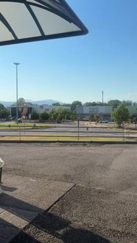 The video shows a daytime outdoor scene with a clear blue sky. In the foreground, there is a section of asphalt paving, with a distinct shadow cast from the left side, indicating an overhang or structure casting the shadow. Moving into the midground, there is a road with several cars driving on it. There are also trees and grassy areas bordering the road. In the background, there are several buildings, which appear to be commercial or retail establishments, with some signage visible. In the far background, mountains or hills are visible against the horizon. The scene is well-lit, suggesting sunny weather.
