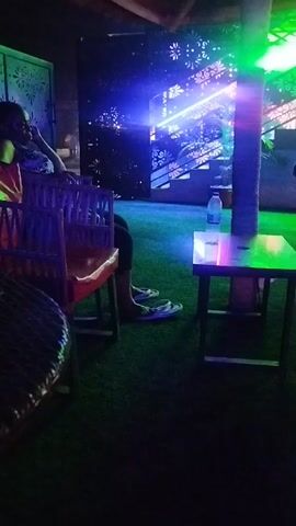 The video shows the interior of what appears to be a bar or lounge. A person is sitting in a wooden chair. They are wearing a red shirt and black pants. Next to them is a small wooden table with a bottle of water on it. The floor is covered in green artificial turf. There is a decorative metal screen behind the person, and stairs lead up behind the screen. Blue and green lights illuminate the area. The overall lighting is dim. Music is playing in the background.