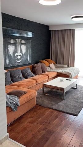 The video shows a brief pan of a well-decorated living room and dining area. The living room has dark hardwood floors, a large orange sectional couch with various throw pillows, and a gray area rug underneath a white coffee table. A large art piece depicting a face hangs on a textured black wall. There are curtains that let in natural light. The view then moves toward a television stand, transitioning to a different flooring, and then a modern white fireplace. Finally, the view rests on a dining area with a white table, pink chairs, a gray rug, and a decorative sculpture. The color scheme is warm with orange, brown, and white tones.