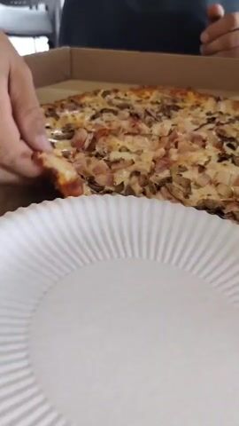 A person's hands are in the frame, grabbing a slice of pizza from a pizza box. The pizza appears to have mushrooms and pieces of meat on it. A white paper plate is in the foreground.