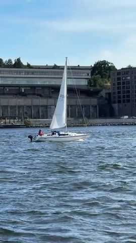 A white sailboat with a single white sail is navigating choppy blue water. Two people are visible on the boat. The boat has the letters "BS" and "CR" visible on its sail. In the background, there is a large, modern-looking building with a long, horizontal facade. Trees are visible behind the building. The sky is blue with some white clouds.