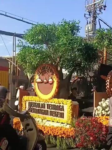 This video shows a parade float decorated with flowers. The float has the words "PT SMELTING" and a slogan about processing tin to build the nation. There are also industrial-looking elements on the float, resembling furnaces and gears. People are visible on and around the float, possibly preparing for or participating in the parade. The environment appears to be outdoors, with trees and buildings in the background.